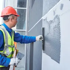 Budowlaniec w kasku i kamizelce wykańcza strukturę elewacyjną, nakładając tynk.