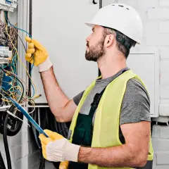 Elektryk w kasku i kamizelce odblaskowej pracuje przy skrzynce z przewodami. Sprawdź nasz cennik usług elektrycznych.
