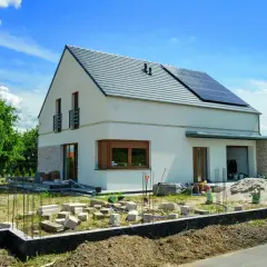 Nowy dom kostka z białym tynkiem i kamienną elewacją, na dachu panele słoneczne. Wokół plac budowy.