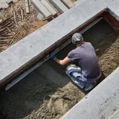 Budowlaniec wyrównuje masę betonową w wykopie. Widać ślady opon na ziemi obok.