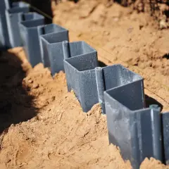 Szare, metalowe elementy, przypominające małe **larseny**, są wkopane w piasek, tworząc linię.