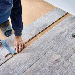 Ręce majsterkowicza mierzą i zaznaczają panele winylowe do cięcia. Młotek i kawałek podłogi leżą obok.