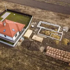 Zabudowa zagrodowa w budowie: gotowy dom z czerwonym dachem obok fundamentów i materiałów budowlanych.