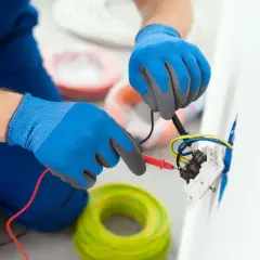 Elektryk w niebieskich rękawiczkach sprawdza kolory kabli w gniazdku. W tle widać zwoje żółto-zielonych przewodów.