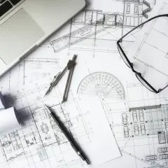 Na biurku leżą plany architektoniczne, laptop, cyrkiel, okulary i długopis. To miejsce pracy nad kosztorysem budowlanym.