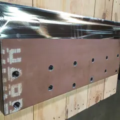 Metalowy element z otworami i oznaczeniami, leżący na drewnianych belkach.