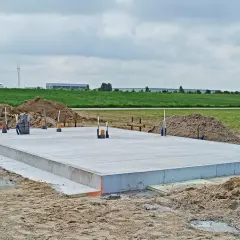 Budowa fundamentów cena: betonowa płyta na placu budowy, otoczona piaskiem i zielenią. W tle widać budynki przemysłowe.