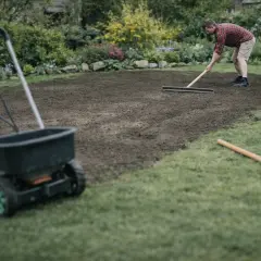 Mężczyzna zgrabia ziemię, przygotowując ją do siewu. W tle widać rozsiewacz do trawy, co sugeruje, że właśnie trwa praca nad tym, jak wyrównać trawnik.