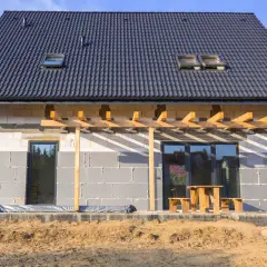 Budowa domu w toku, ściany z bloczków betonowych czekają na ocieplenie domu styropianem. Dach pokryty dachówką, widoczne okna dachowe i tarasowe.