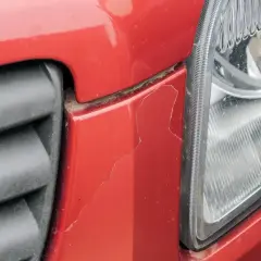 Pęknięty lakier na czerwonym elemencie samochodu przy grillu i reflektorze. Sprawdź lakierowanie elementu samochodu cennik.