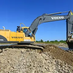 Żółta koparka Volvo EC240B LC pracuje na kamienistym brzegu rzeki. Zastanawiasz się, ile kosztuje godzina pracy koparki?