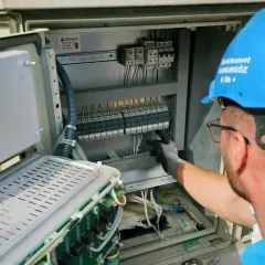 Elektryk w niebieskim kasku pracuje przy panelu sterowania. Potwierdza gotowość instalacji przyłączanej, dbając o każdy szczegół.