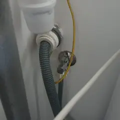 Szara rura odpływowa pralki podłączona do syfonu pod zlewem. Widoczny żółty przewód i biały kabel.