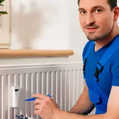 Fachowiec w niebieskim kombinezonie sprawdza odczyty na podzielnikach ciepła zamontowanych na grzejniku.