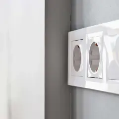 Białe gniazdka elektryczne w ramce na szarej ścianie.