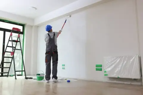 Malowanie ścian w mieszkaniu 50 m. Pracownik w kasku i ogrodniczkach maluje wałkiem. Obok drabina i wiadro z farbą.