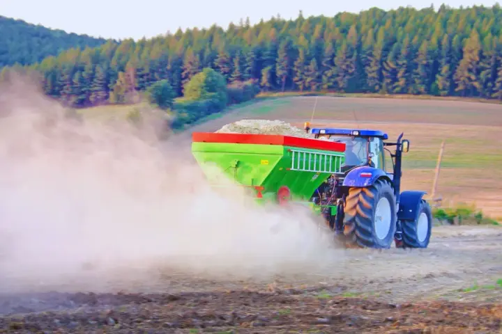 Traktor z zielonym rozsiewaczem rozsypuje wapno, pomagając w odkwaszaniu gleby. W tle las i pole.