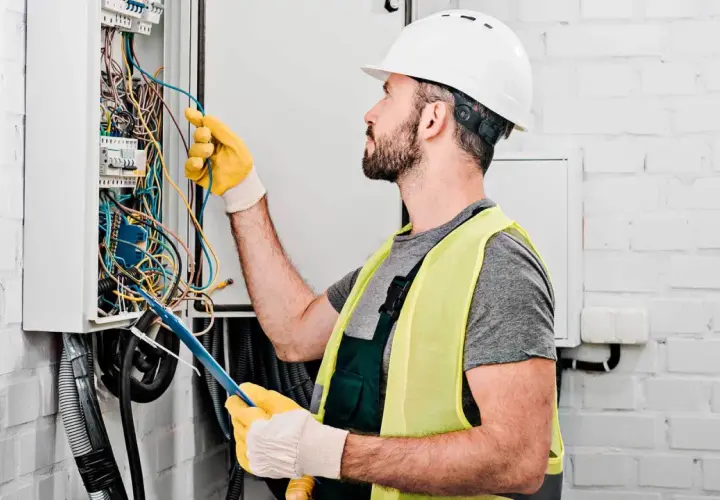 Elektryk w kasku i kamizelce odblaskowej pracuje przy skrzynce z przewodami. Sprawdź nasz cennik usług elektrycznych.