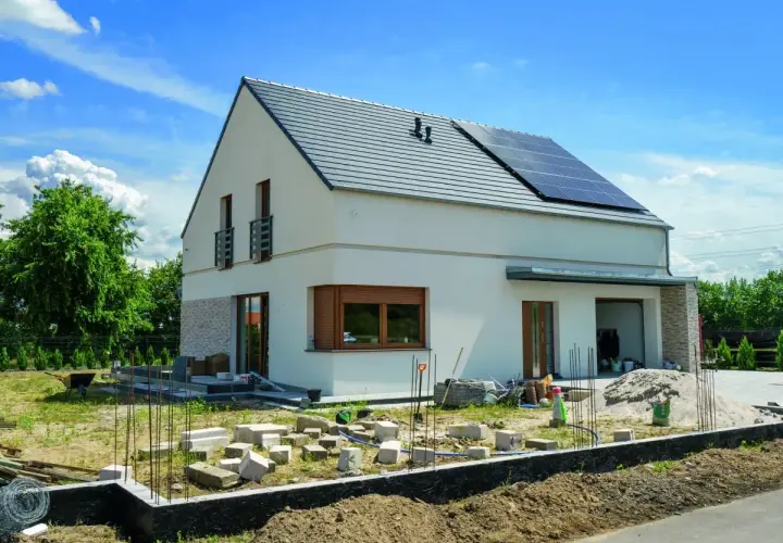 Nowy dom kostka z białym tynkiem i kamienną elewacją, na dachu panele słoneczne. Wokół plac budowy.