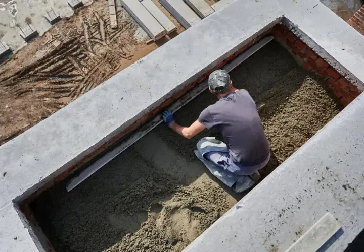 Budowlaniec wyrównuje masę betonową w wykopie. Widać ślady opon na ziemi obok.