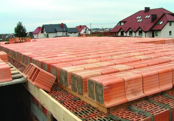 Strop ackermana w budowie, rzędy pustaków ceramicznych czekają na betonowanie. W tle domy z czerwonymi dachami.