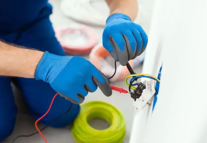 Elektryk w niebieskich rękawiczkach sprawdza kolory kabli w gniazdku. W tle widać zwoje żółto-zielonych przewodów.