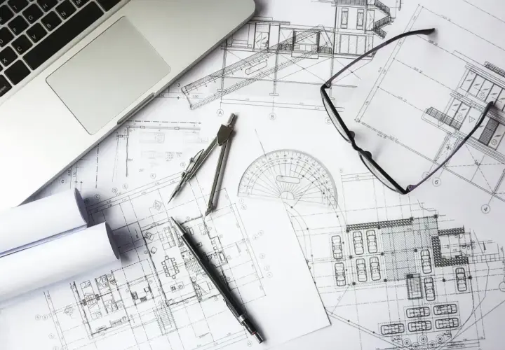 Na biurku leżą plany architektoniczne, laptop, cyrkiel, okulary i długopis. To miejsce pracy nad kosztorysem budowlanym.