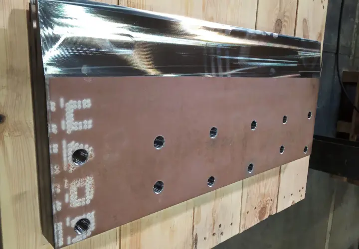 Metalowy element z otworami i oznaczeniami, leżący na drewnianych belkach.