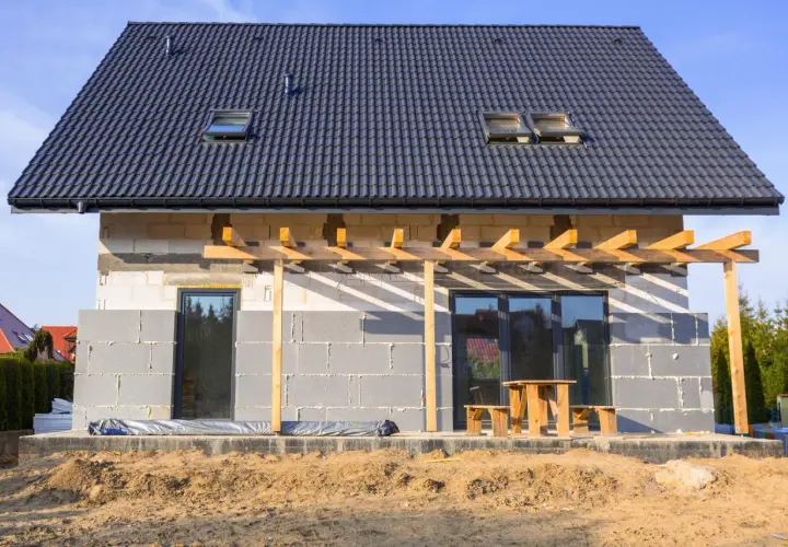Budowa domu w toku, ściany z bloczków betonowych czekają na ocieplenie domu styropianem. Dach pokryty dachówką, widoczne okna dachowe i tarasowe.