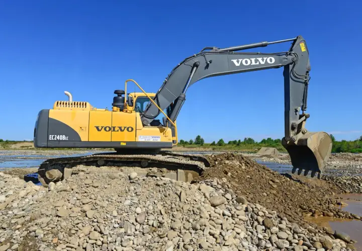 Żółta koparka Volvo EC240B LC pracuje na kamienistym brzegu rzeki. Zastanawiasz się, ile kosztuje godzina pracy koparki?