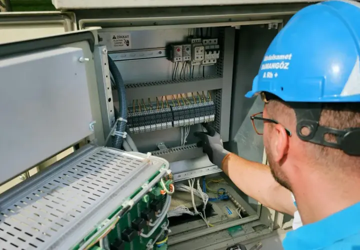 Elektryk w niebieskim kasku pracuje przy panelu sterowania. Potwierdza gotowość instalacji przyłączanej, dbając o każdy szczegół.