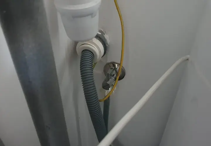 Szara rura odpływowa pralki podłączona do syfonu pod zlewem. Widoczny żółty przewód i biały kabel.