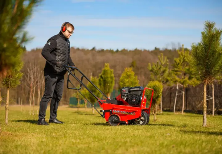 Mężczyzna w słuchawkach i okularach prowadzi czerwoną aerator. Idealny czas na aerację trawnika, by zapewnić mu zdrowy wzrost.