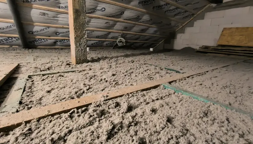 Izolacja poddasza z celulozy, która to materiał ekologiczny i skuteczny. Drewniane belki i folia dachowa widoczne nad warstwą izolacji.