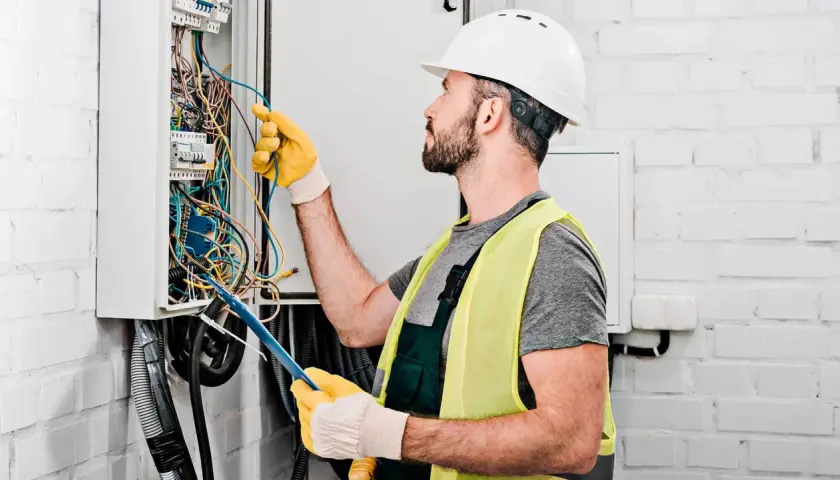Elektryk w kasku i kamizelce odblaskowej pracuje przy skrzynce z przewodami. Sprawdź nasz cennik usług elektrycznych.