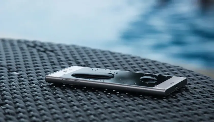 Telefon z kroplami wody na ekranie, leżący na plecionej powierzchni. IP65 co oznacza? Ochrona przed pyłem i strumieniem wody.