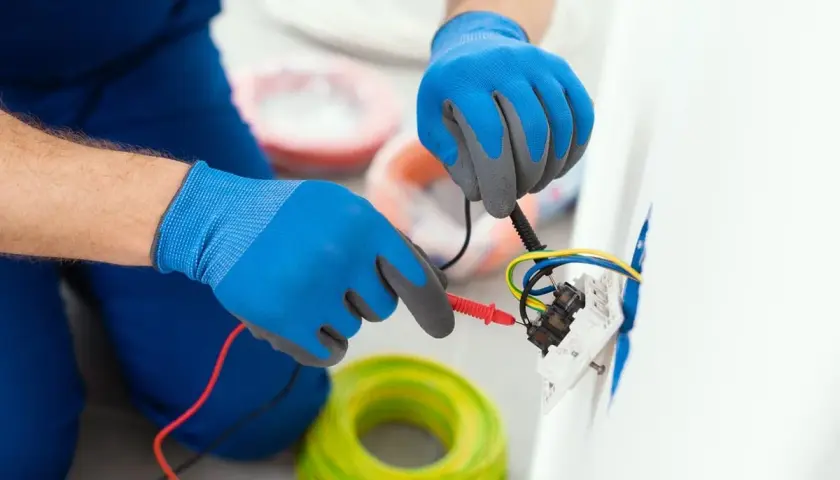 Elektryk w niebieskich rękawiczkach sprawdza kolory kabli w gniazdku. W tle widać zwoje żółto-zielonych przewodów.