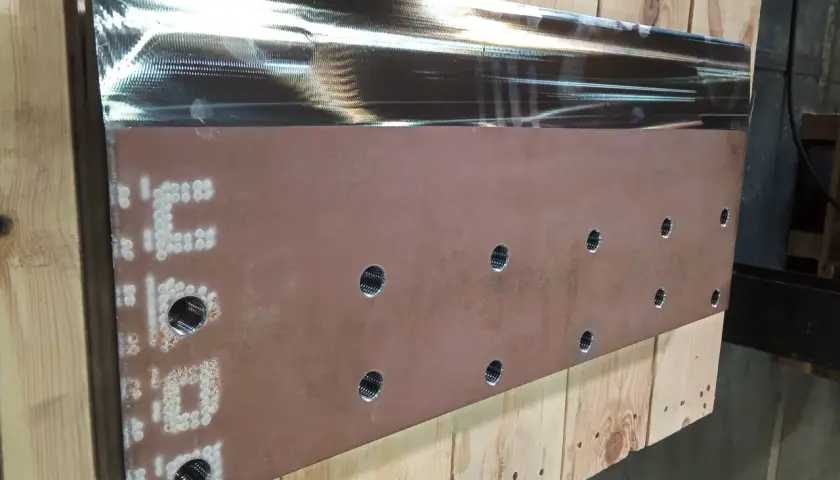Metalowy element z otworami i oznaczeniami, leżący na drewnianych belkach.