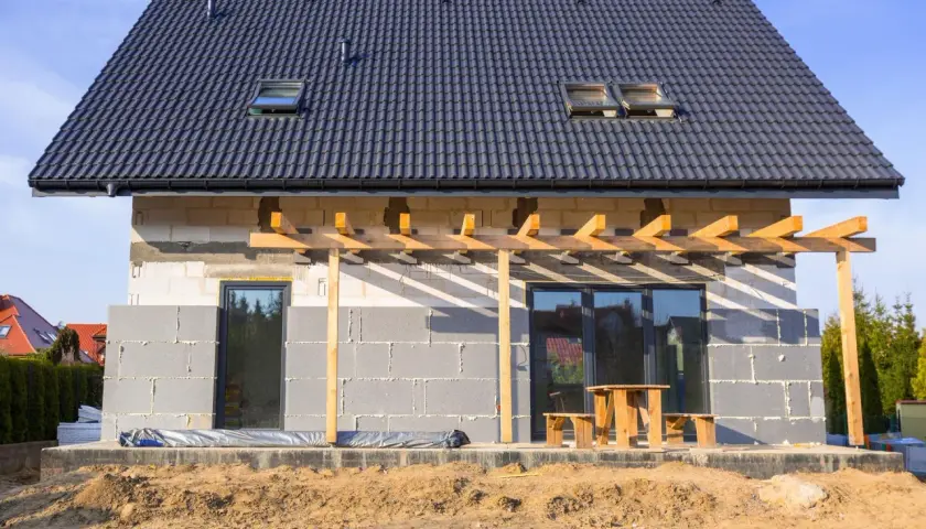 Budowa domu w toku, ściany z bloczków betonowych czekają na ocieplenie domu styropianem. Dach pokryty dachówką, widoczne okna dachowe i tarasowe.