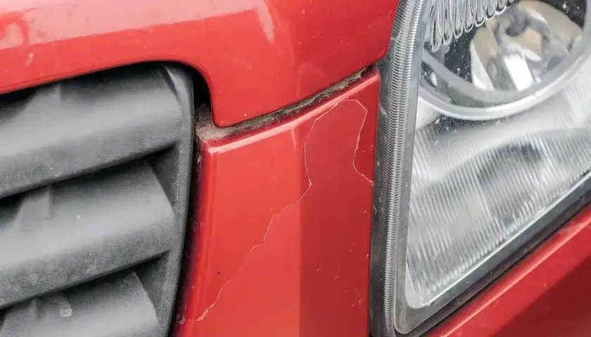 Pęknięty lakier na czerwonym elemencie samochodu przy grillu i reflektorze. Sprawdź lakierowanie elementu samochodu cennik.
