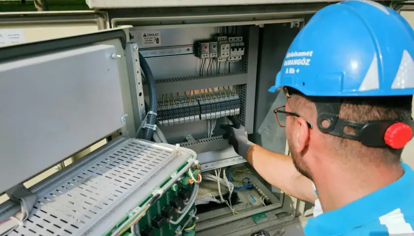Elektryk w niebieskim kasku pracuje przy panelu sterowania. Potwierdza gotowość instalacji przyłączanej, dbając o każdy szczegół.