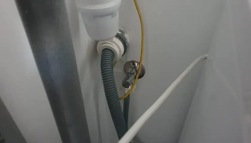 Szara rura odpływowa pralki podłączona do syfonu pod zlewem. Widoczny żółty przewód i biały kabel.