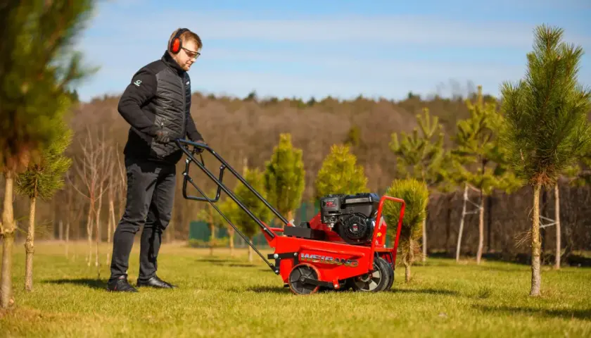 Mężczyzna w słuchawkach i okularach prowadzi czerwoną aerator. Idealny czas na aerację trawnika, by zapewnić mu zdrowy wzrost.