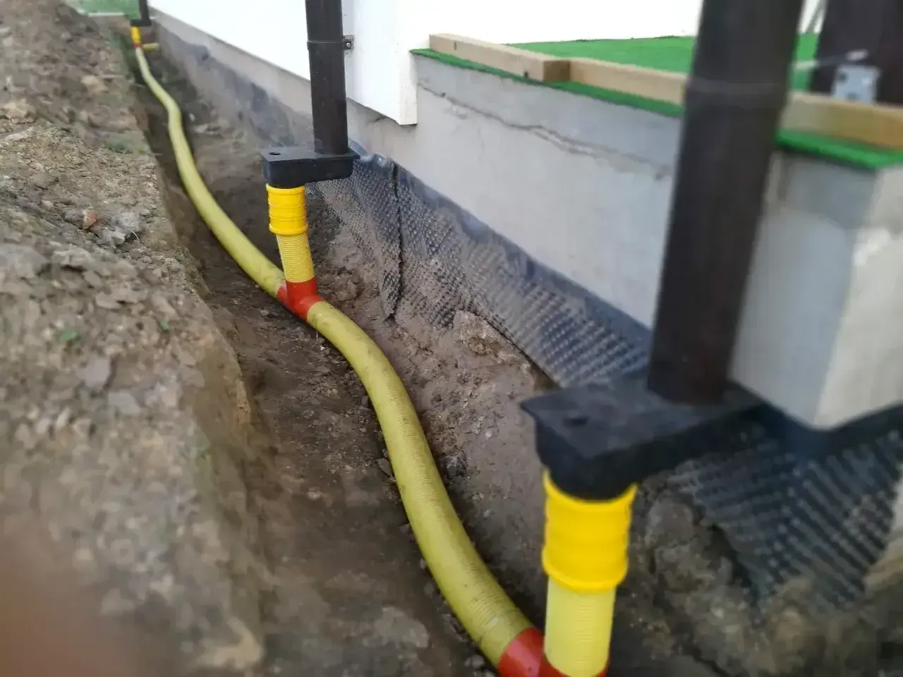 System odprowadzenia wody z rynien w żółtej rurze, biegnącej w wykopie przy fundamencie domu.