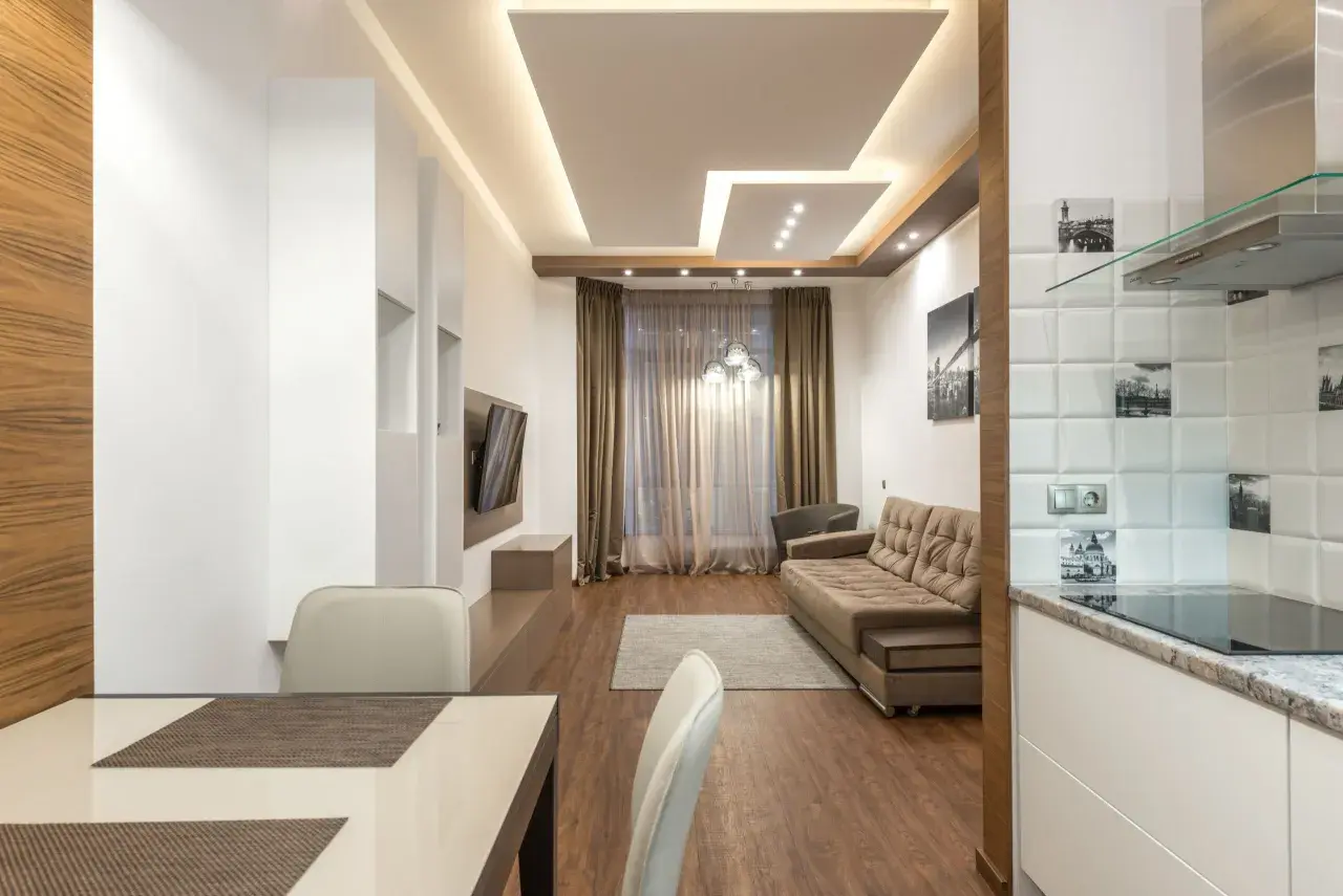Nowoczesny salon z designerskim sufitem podwieszanym, kanapą i aneksem kuchennym.