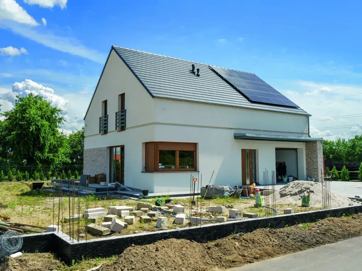 Nowy dom kostka z białym tynkiem i kamienną elewacją, na dachu panele słoneczne. Wokół plac budowy.