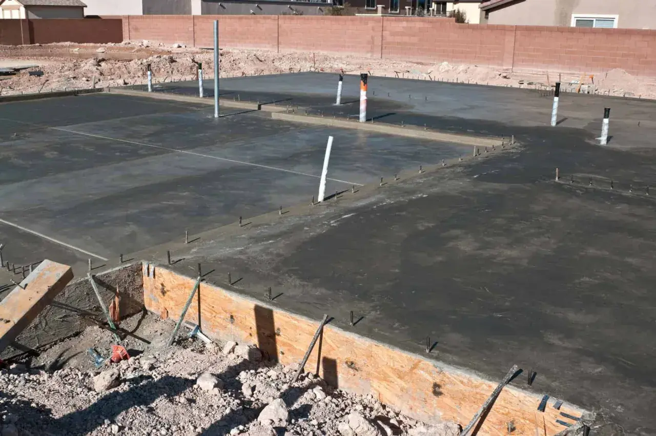 Świeżo wylany beton na płycie fundamentowej, widoczne rury i zbrojenie. Trwa izolacja pozioma fundamentów.