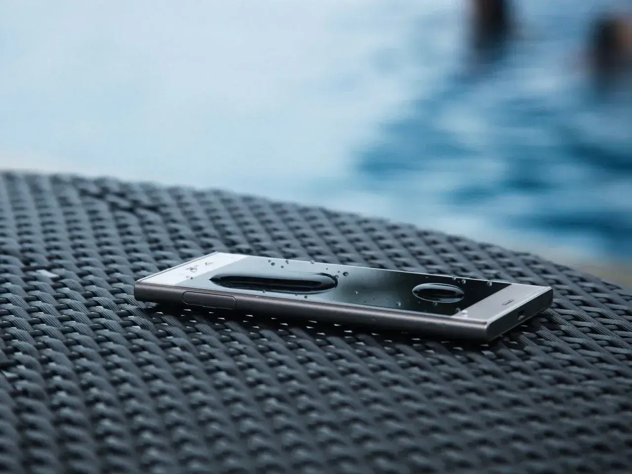 Telefon z kroplami wody na ekranie, leżący na plecionej powierzchni. IP65 co oznacza? Ochrona przed pyłem i strumieniem wody.