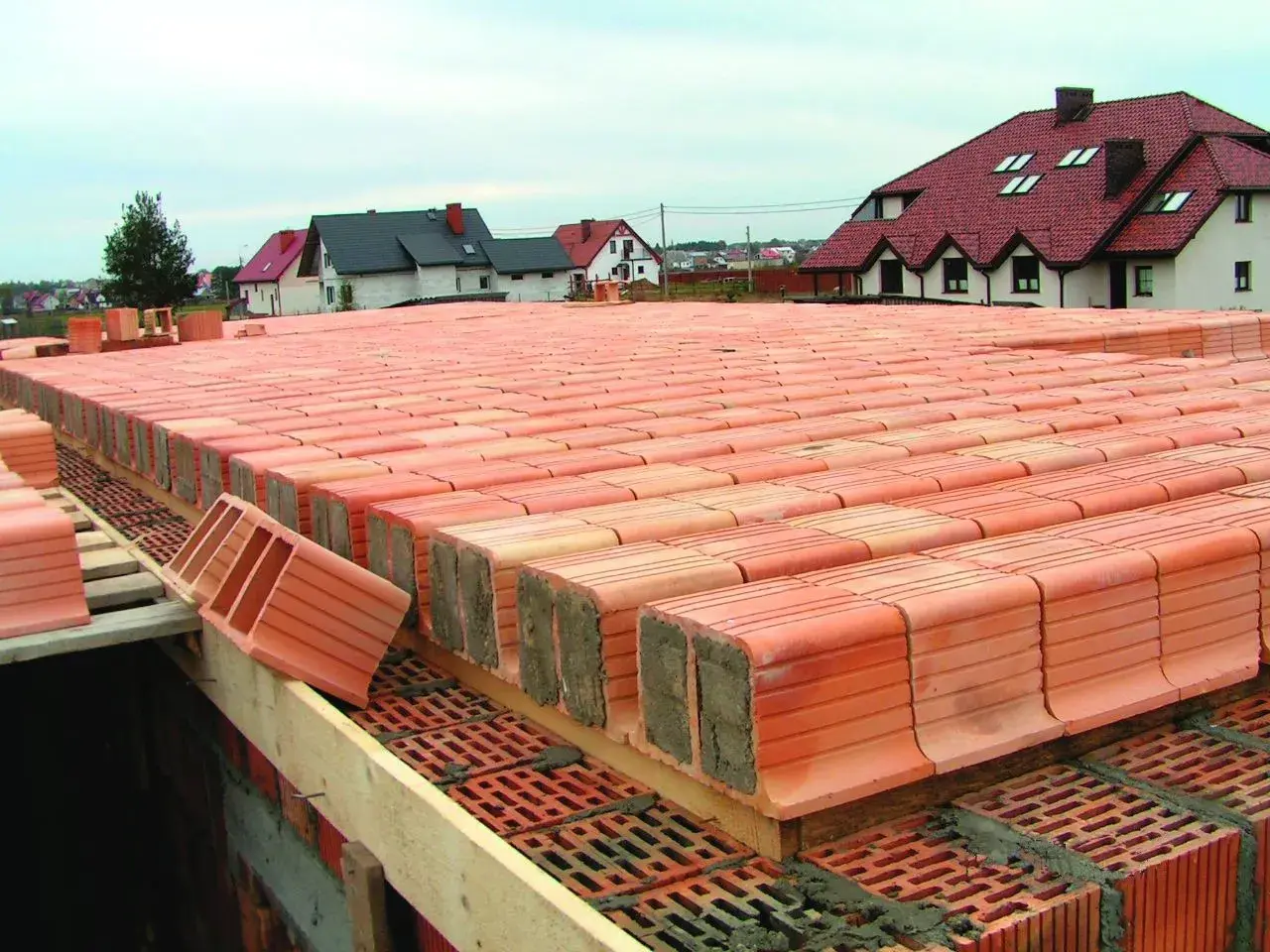 Strop ackermana w budowie, rzędy pustaków ceramicznych czekają na betonowanie. W tle domy z czerwonymi dachami.