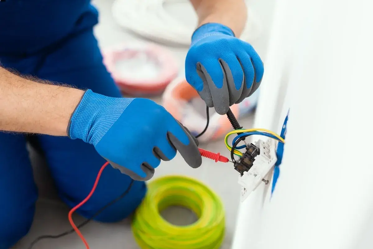 Elektryk w niebieskich rękawiczkach sprawdza kolory kabli w gniazdku. W tle widać zwoje żółto-zielonych przewodów.